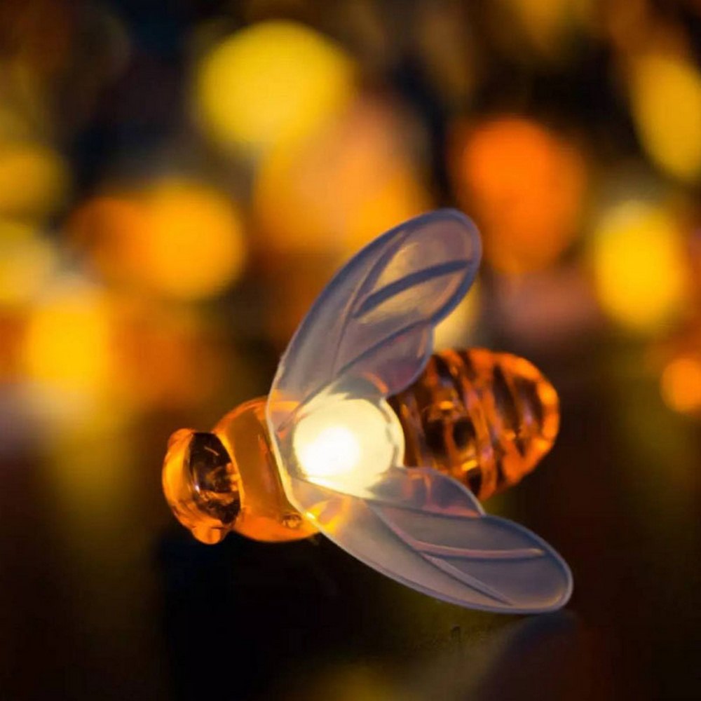 Close-up of a glowing bee-shaped light decoration with translucent wings, set against a blurred background of warm, yellow-orange bokeh lights.