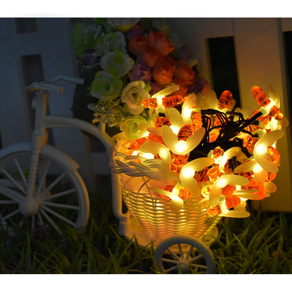A white decorative bicycle holds a basket of colorful artificial flowers and glowing bee-shaped string lights, creating a warm, inviting ambiance against a dark backdrop.