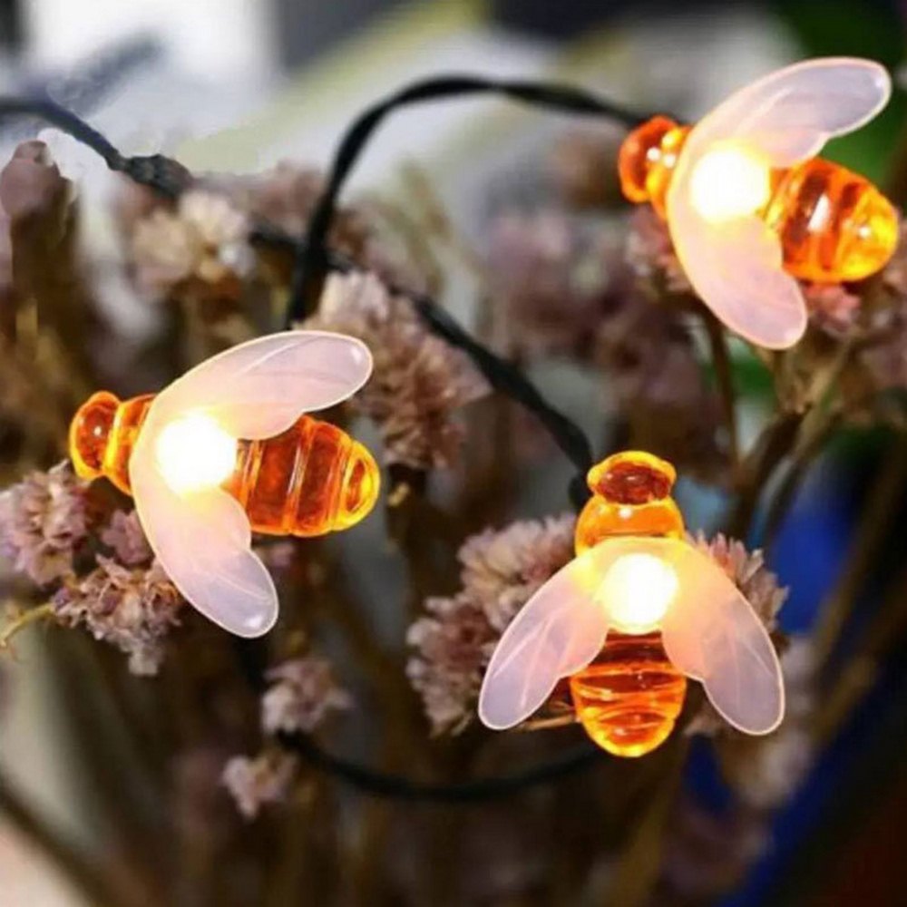String lights shaped like bees with glowing orange bodies and translucent wings, draped over dried flowers. The soft illumination creates a warm, whimsical ambiance.