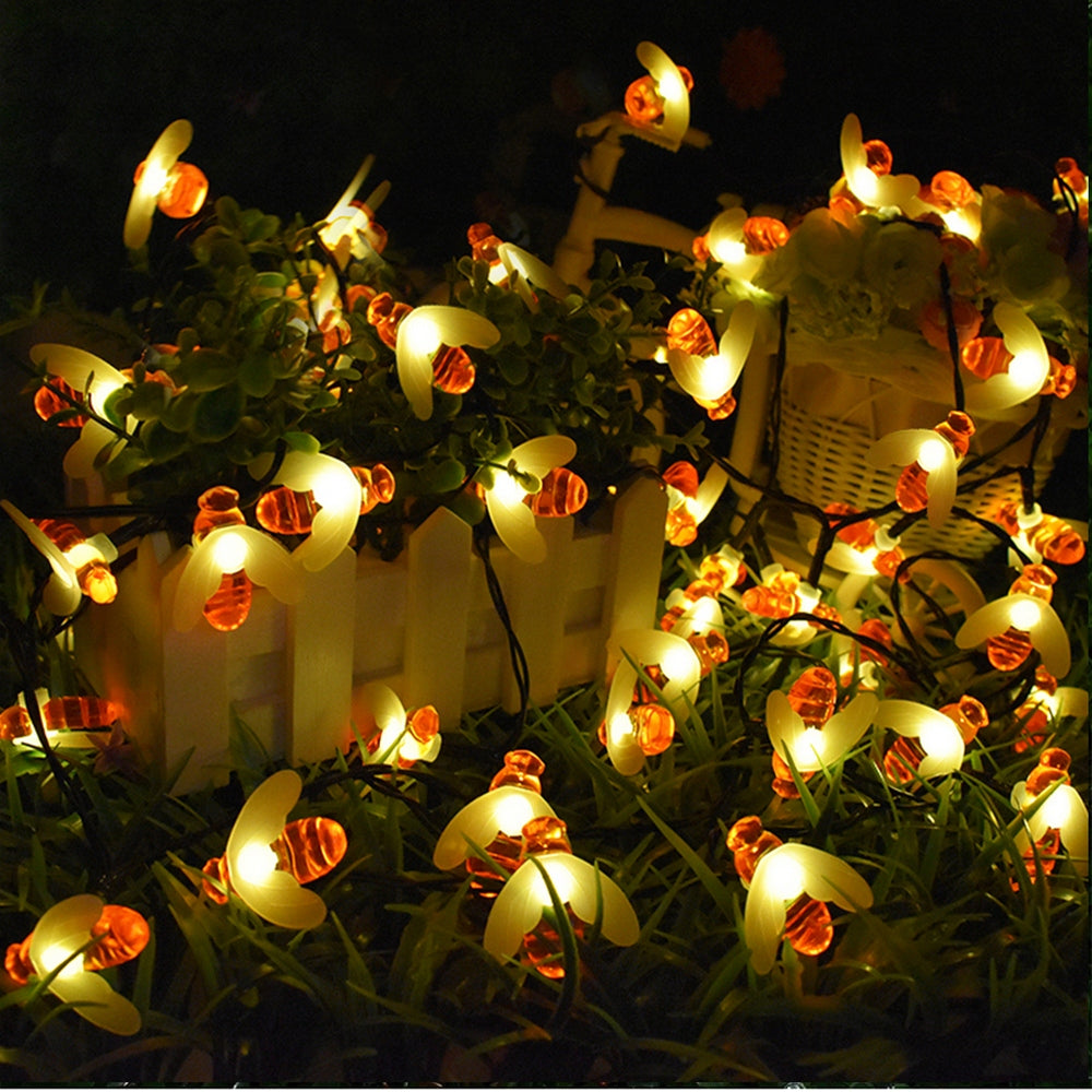 Glowing bee-shaped string lights draped over plants and white picket fence in a dark setting, creating a warm, whimsical ambiance.