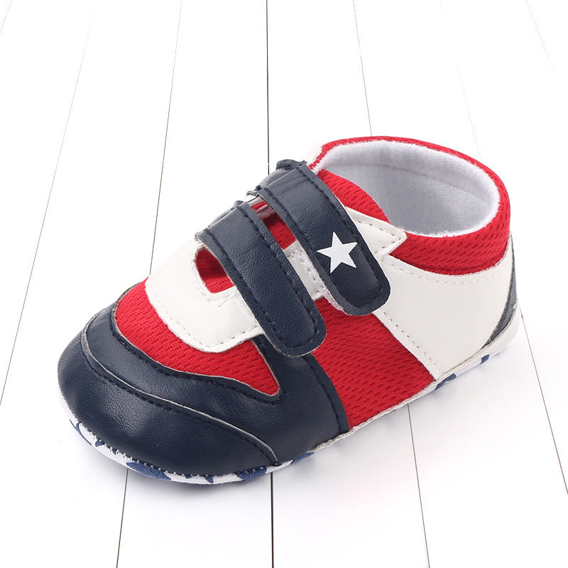 Red, white, and navy blue baby shoe with a star design. The shoe features two velcro straps and is displayed on a white wooden surface.