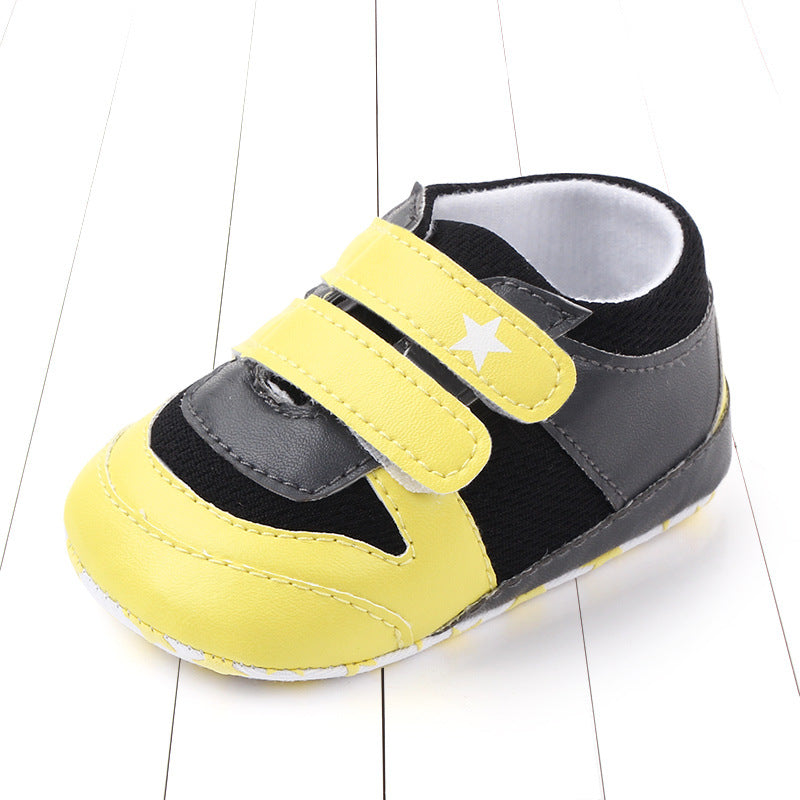 Yellow and black baby sneaker with two velcro straps and a white star design on one strap, displayed on a white wood background.