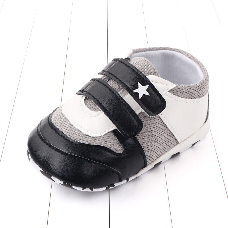 Stylish baby shoe with black and white design, featuring a star on the black velcro strap and a mesh gray section, placed on a wooden floor with a white panel pattern.