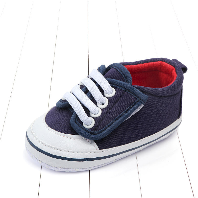 Navy blue and white baby sneaker with white laces and red interior, displayed on a white wooden surface. Features a thick sole and a pull tab at the back.