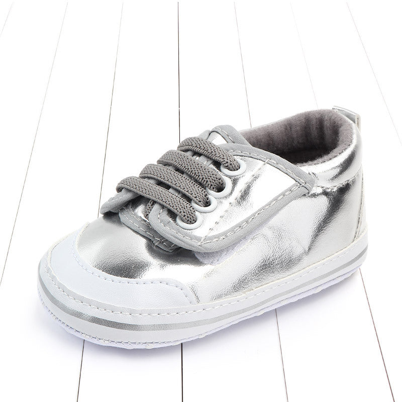 Shiny silver baby sneaker with gray laces and white accents, placed on a white wooden surface.