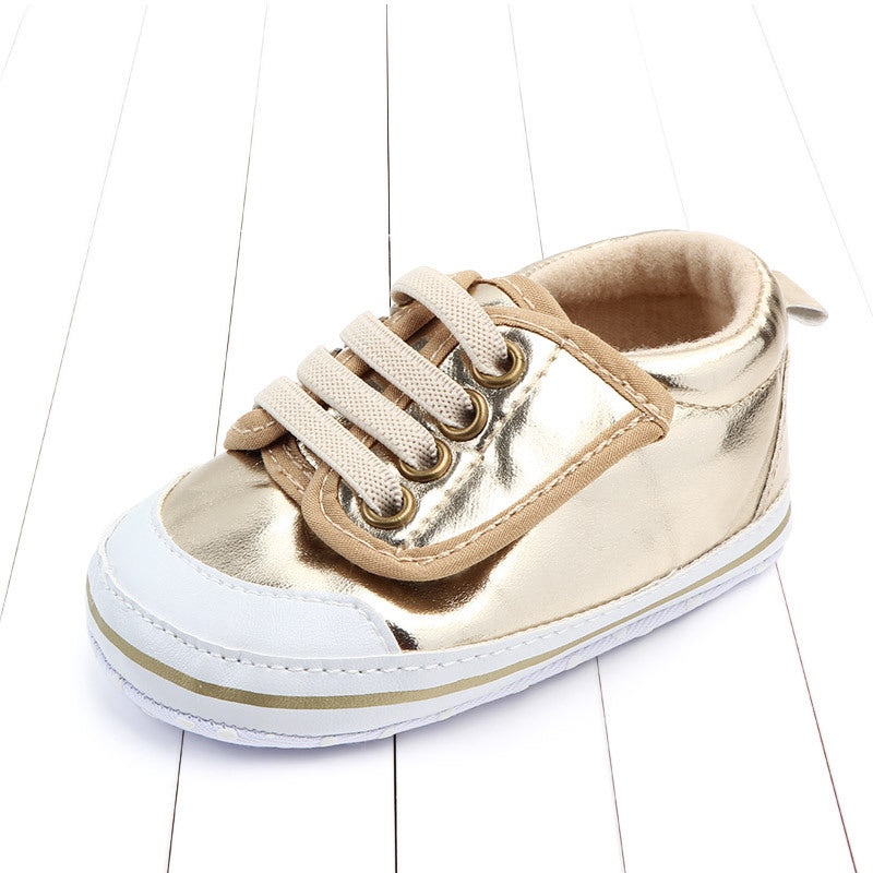 A shiny gold baby sneaker with beige laces, white toe cap, and gold striped sole, placed on a white wooden floor.