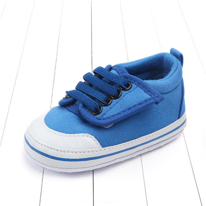 Blue toddler sneaker with black laces on a white plank floor. The shoe has a white sole with a blue stripe, featuring a fabric upper and reinforced white toe cap.