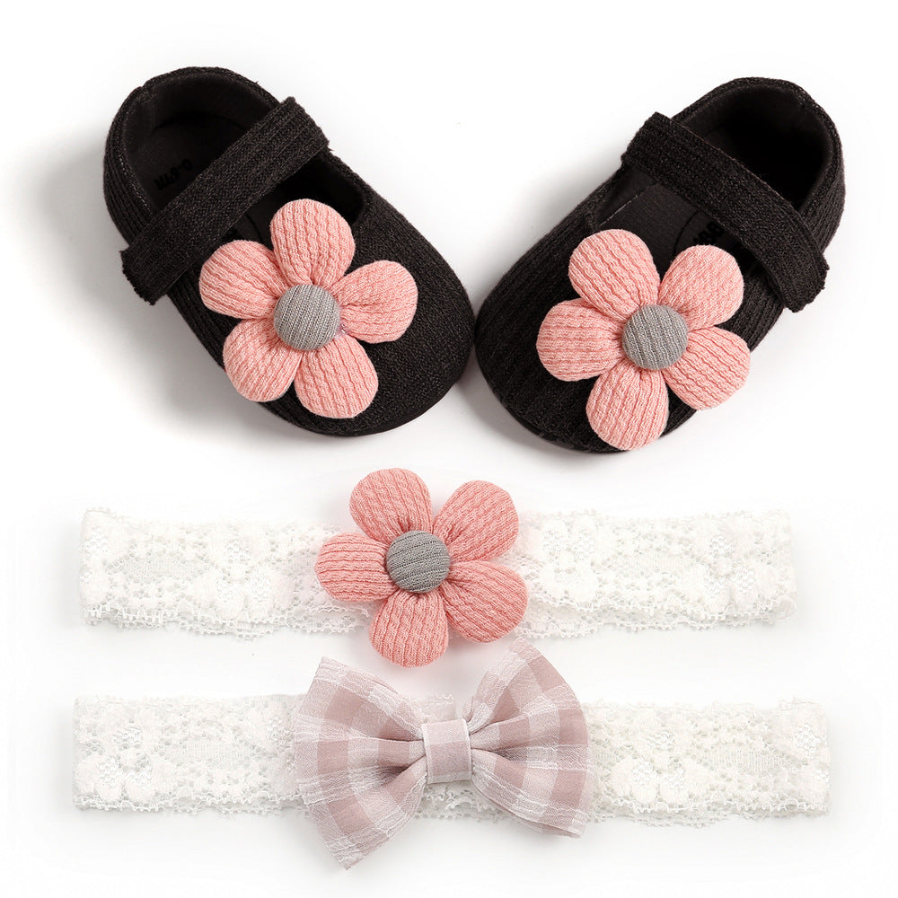 Black baby shoes with pink flower accents, paired with two lace headbands—one with a pink flower and the other with a checkered bow, on a white background.