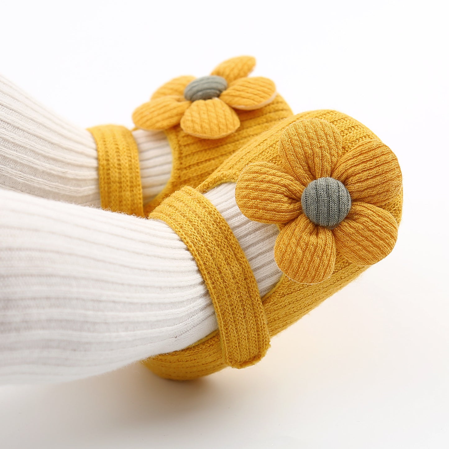 Yellow knit baby shoes adorned with large flower appliques. The shoes have ankle straps, and a child's legs wearing white ribbed pants are visible. The background is white.