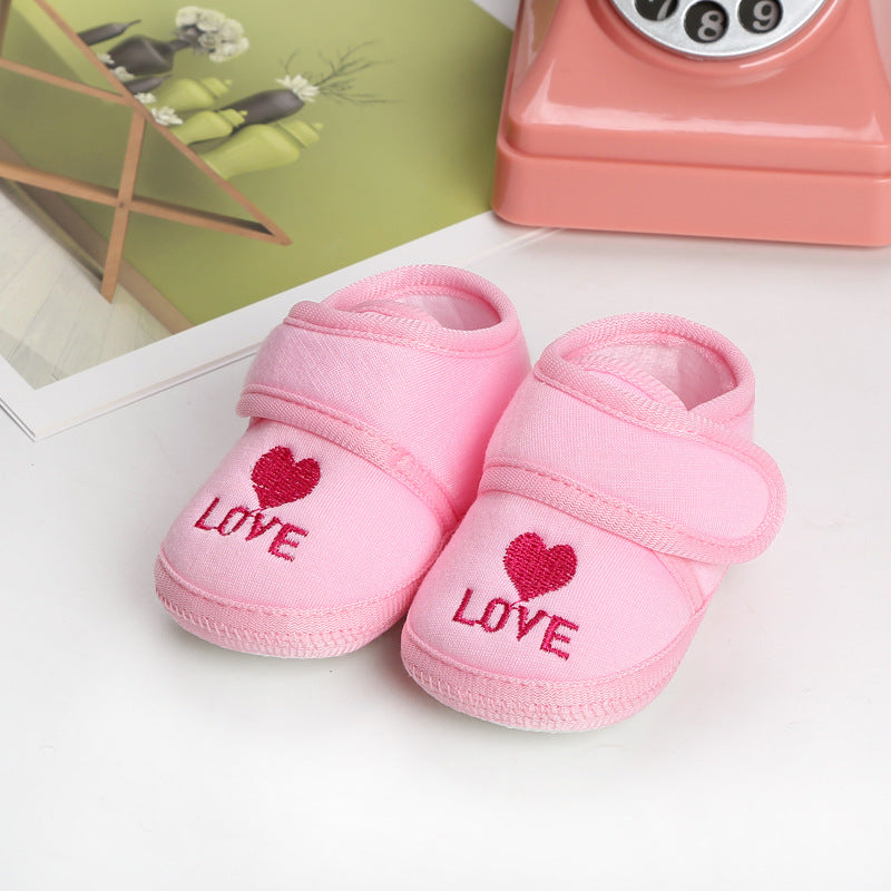Pink baby shoes with "LOVE" and a heart embroidered in red. They're placed beside a pink rotary phone and a modern green vase on a light surface.