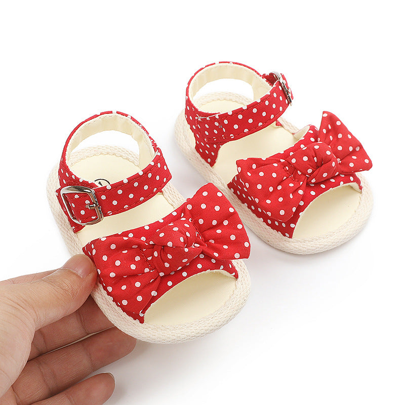 Red polka dot baby sandals with bows and ankle straps. One sandal is held in a hand, showing a silver buckle. Soft soles and open toes on a white background.