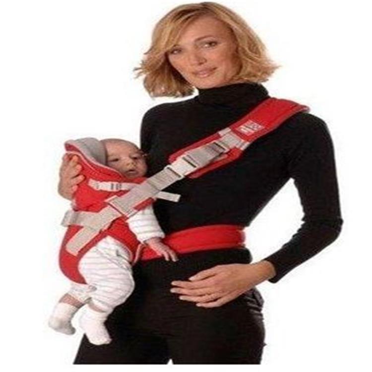 A woman in a black turtleneck carries a baby in a red and gray baby carrier. The baby is wearing a striped outfit and appears content, while the woman smiles at the camera.