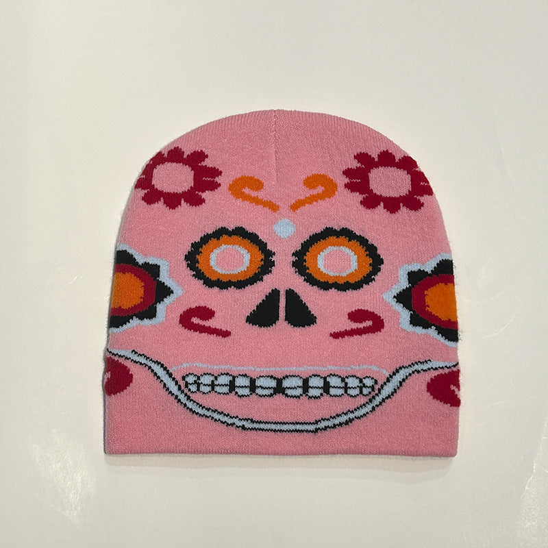 A pink knit beanie featuring a colorful sugar skull design with floral patterns, orange eyes, and a grin, reminiscent of Dia de los Muertos motifs, on a plain background.