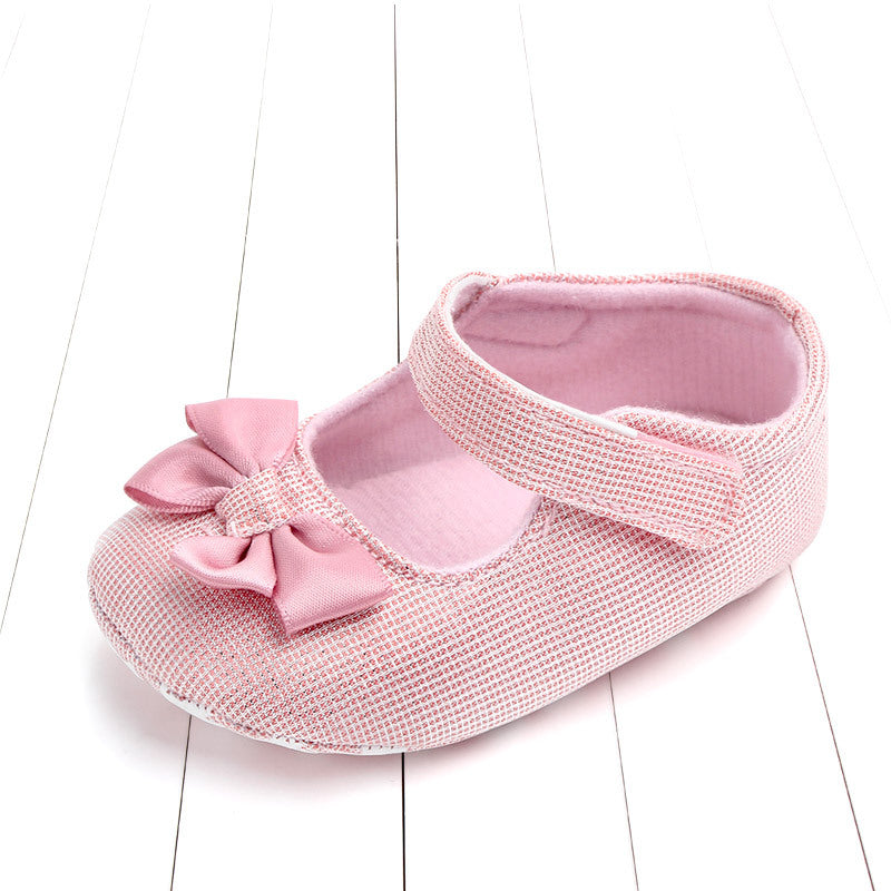 Pink baby shoe with a textured pattern, featuring a Velcro strap and a decorative bow on the front, placed on a white wooden background.