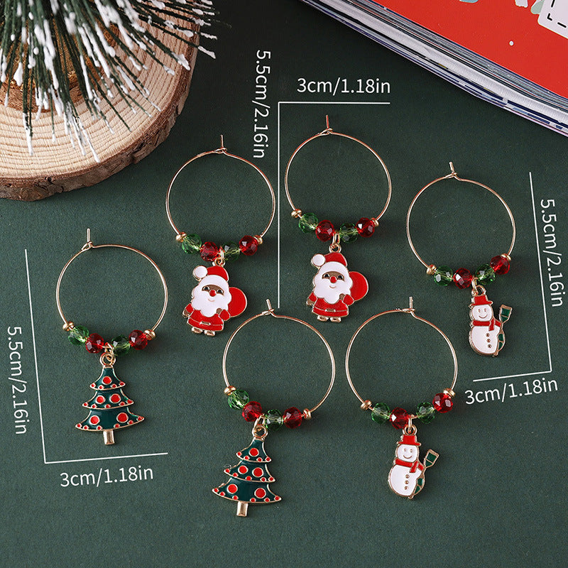 Six festive wine glass charms featuring Santa, snowmen, and Christmas trees on gold rings with red and green beads. They're displayed on a dark green surface next to a pine branch.