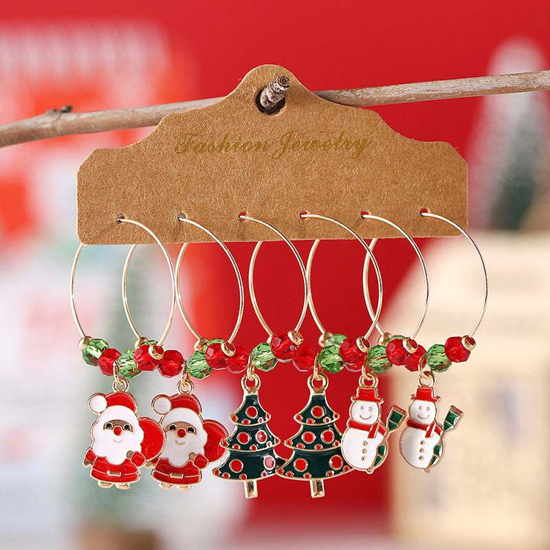 Festive wine glass charms with Santa, Christmas tree, and snowman designs, adorned with red and green beads, displayed on a card against a blurred red background.