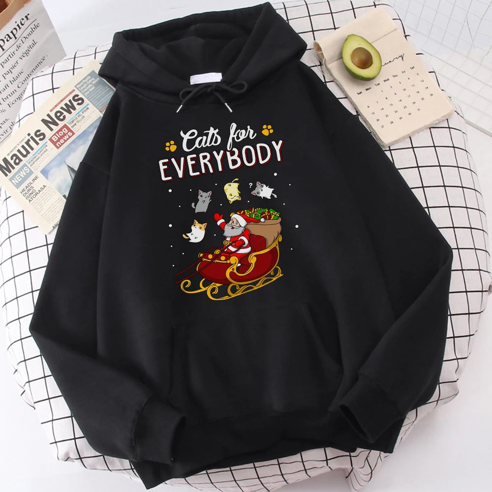 Black hoodie with a festive design featuring a cartoon Santa in a sleigh surrounded by cats. Text reads "Cats for Everybody." Background shows a grid pattern, newspaper, and avocado.