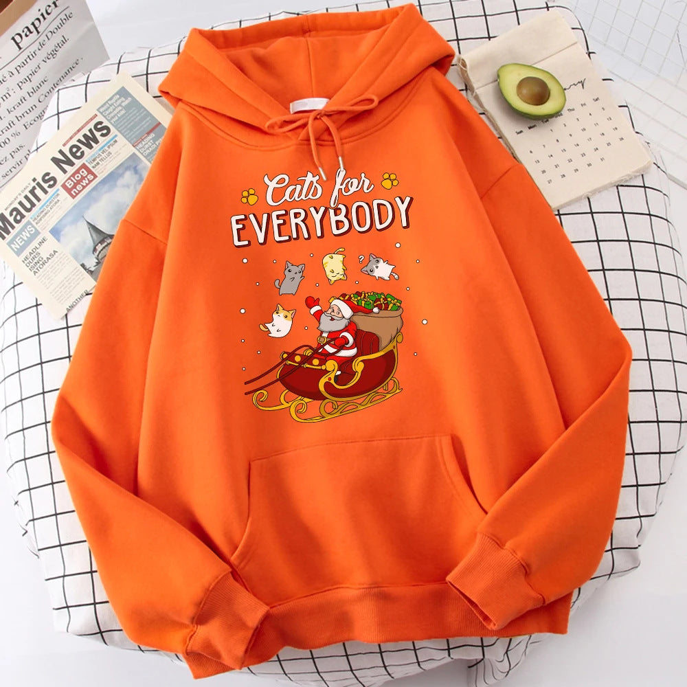 Orange hoodie with a Santa cat design in a sleigh surrounded by floating cats. Text reads "Cats for Everybody." Background features a newspaper, notepad, and halved avocado.