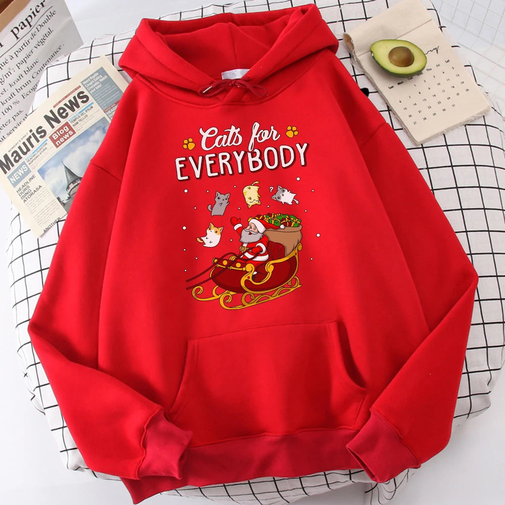 Red hoodie with "Cats for Everybody" text and a graphic of a Santa cat in a sleigh surrounded by playful cats. It lies on a checkered surface beside a newspaper and half an avocado.