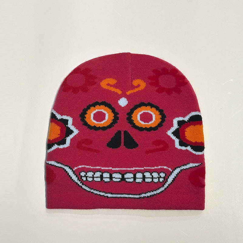 Red beanie with colorful Day of the Dead skull design. Features orange and black eyes, floral elements, and a wide, white-toothed smile on a soft fabric.