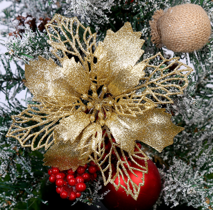 Golden poinsettia decoration with glittery leaves on a snow-dusted Christmas tree, accompanied by red berries and a red bauble, with a burlap-wrapped ornament nearby.