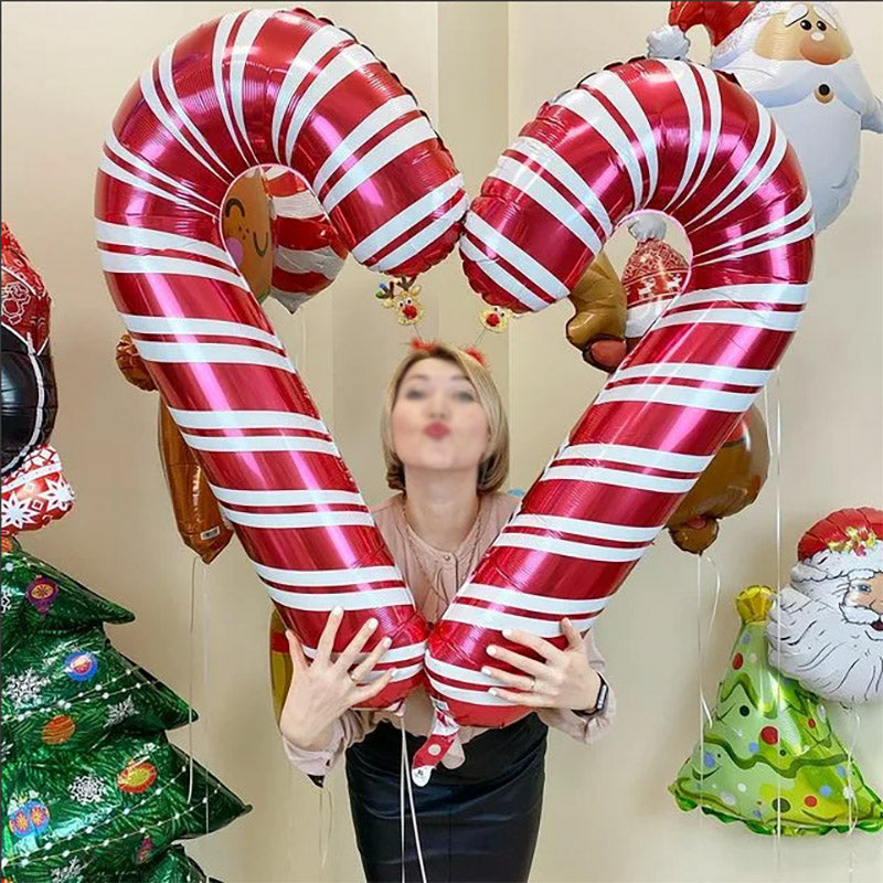 A woman holds two large candy cane balloons forming a heart shape. She wears reindeer antlers and stands among festive balloons, including a Christmas tree and Santa Claus.