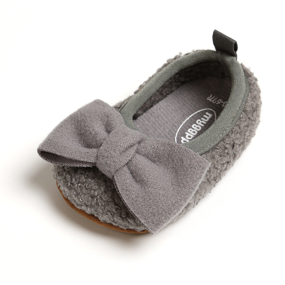 A cozy, gray baby shoe with a fluffy texture and a large, soft bow on top. The shoe features a soft lining and a small pull tab at the heel.