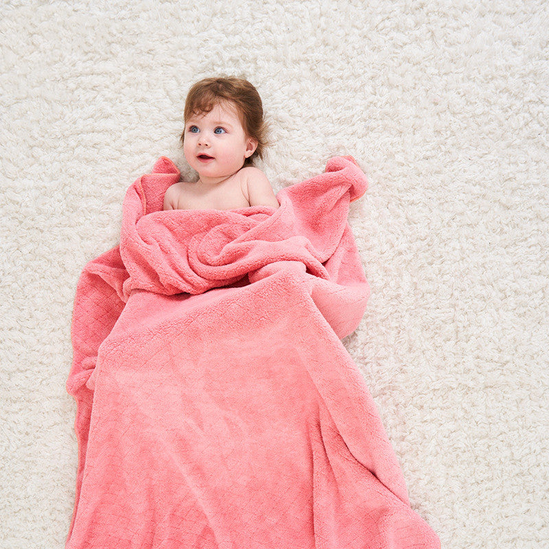 A baby with reddish-brown hair is wrapped in a soft pink blanket, lying on a fluffy, light-colored carpet, looking up with a curious expression.