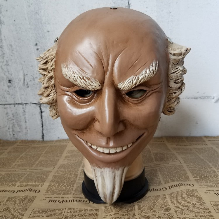 A sculpted mask with a mischievous grin, sharp eyebrows, narrow eyes, and white, curly sideburns. The mask has a small goatee and sits on a newspaper-covered surface.