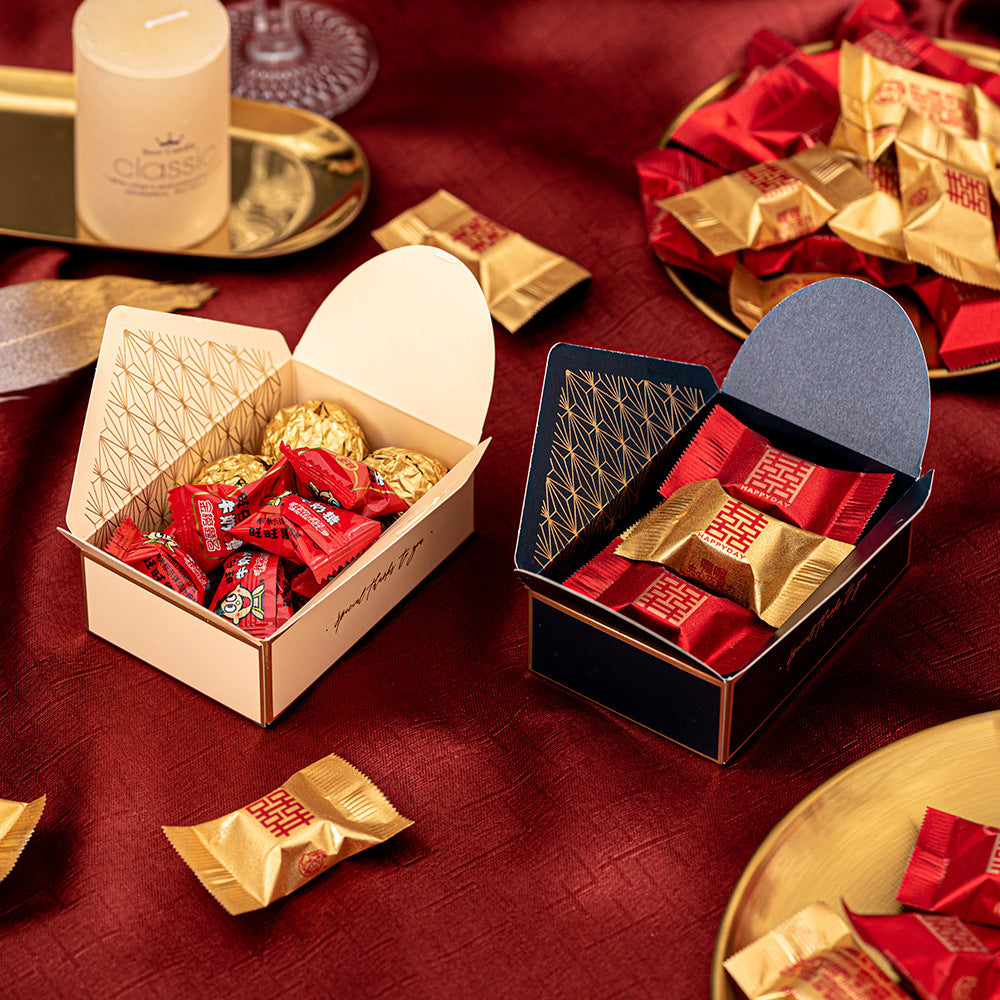 Two decorative boxes with assorted candies, one box open to show gold-wrapped chocolates and red sweets, placed on a red cloth with additional candies scattered around.