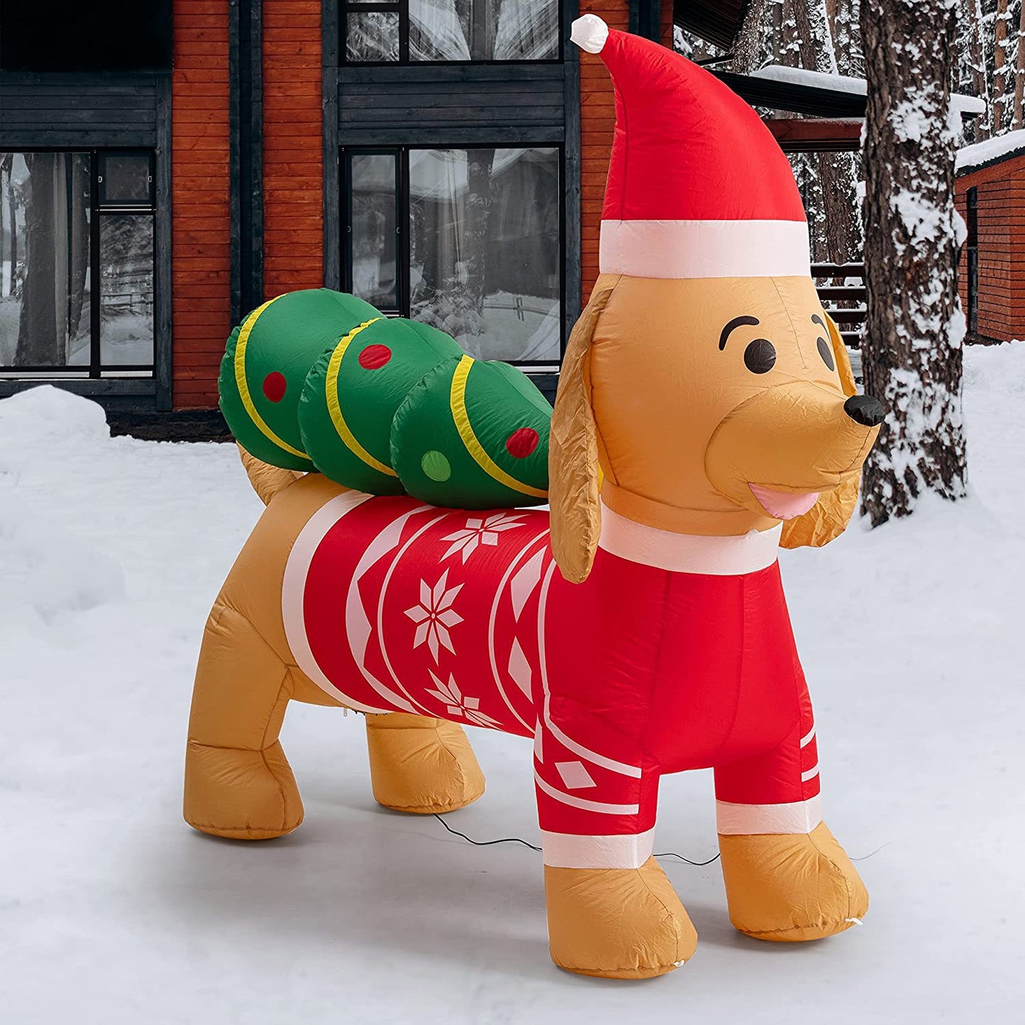 Inflatable dog in a Santa hat and red sweater with snowflakes, carrying a decorated green Christmas tree on its back, set outside a snow-covered wooden house.