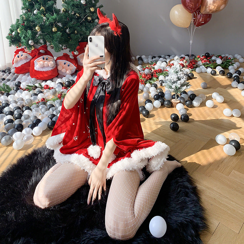 Woman in a red Santa dress with devil horns takes a selfie. She sits on a faux fur rug with white stockings. Christmas tree and festive decor, including Santa sacks, are in the background.