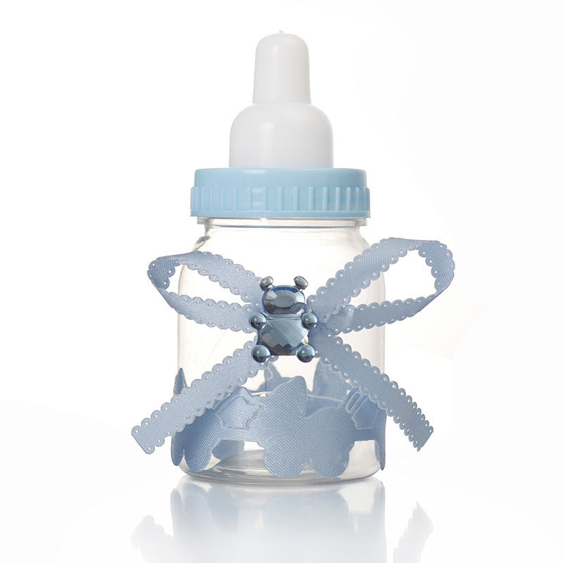 A clear plastic baby bottle with a white nipple and blue cap. It is adorned with a decorative blue ribbon and a jewel-like embellishment, alongside paper decorations inside.