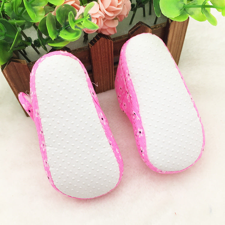 Pink baby shoes with white soles and small floral patterns are placed upside down on a soft surface, surrounded by green leaves and pink flowers in a wooden container.