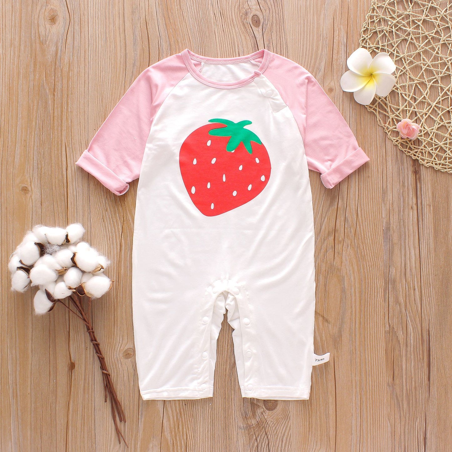 White baby romper with pink sleeves and a large red strawberry print on the front, placed on a wooden surface with cotton sprigs and flowers.
