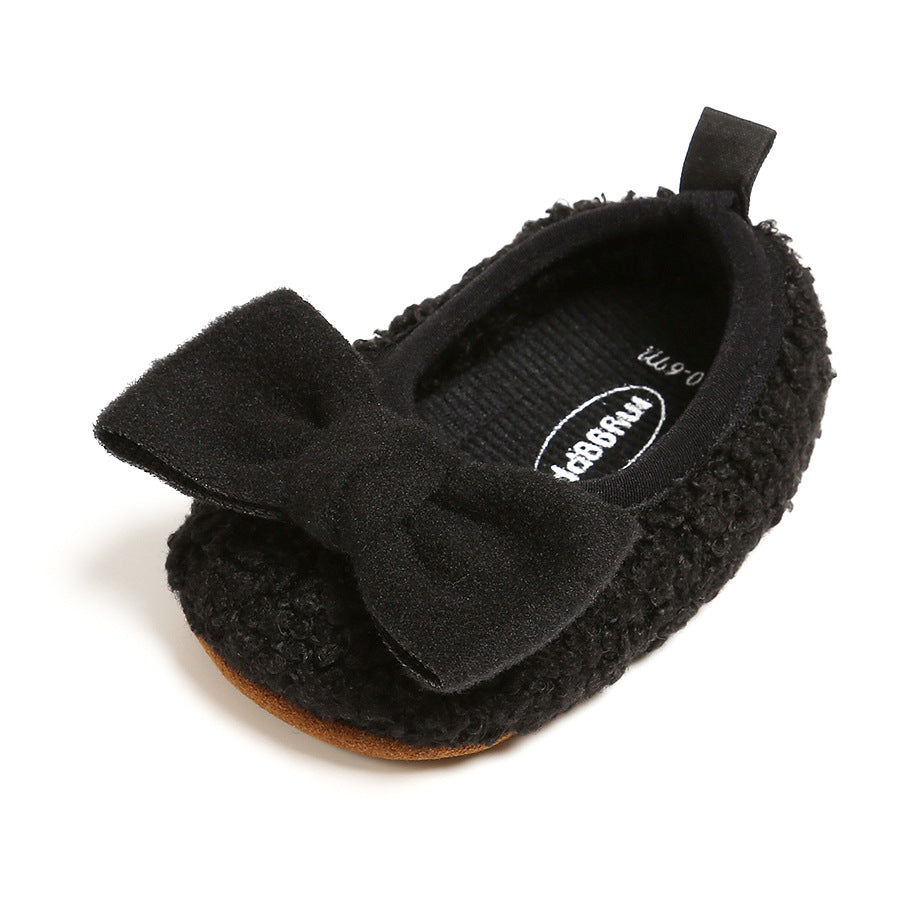 A stylish black baby shoe with a textured surface, featuring a soft fabric bow on top. The shoe has a small pull tab at the back and a contrasting brown sole.