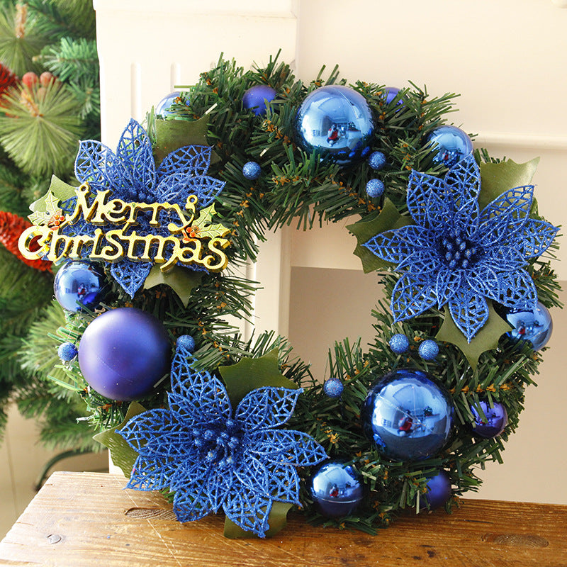 Festive Christmas wreath adorned with blue ornaments, lace-like blue poinsettias, and a "Merry Christmas" sign, placed on a wooden table near greenery.