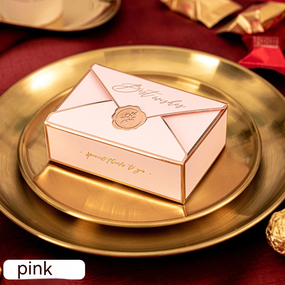 Pink envelope-shaped box labeled "Best wishes" rests on a gold plate. The background features folded red and gold paper decorations.