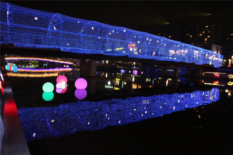 Bridge adorned with blue lights reflecting on water at night. Colorful glowing orbs float on the river, and vivid reflections create a mesmerizing visual display.