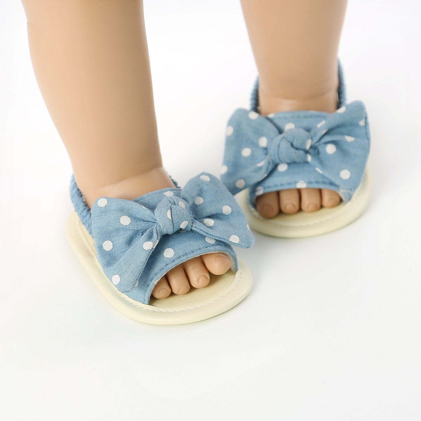 Baby feet wearing blue polka dot sandals with large bows. The sandals are open-toed and have stretchy fabric around the ankles.