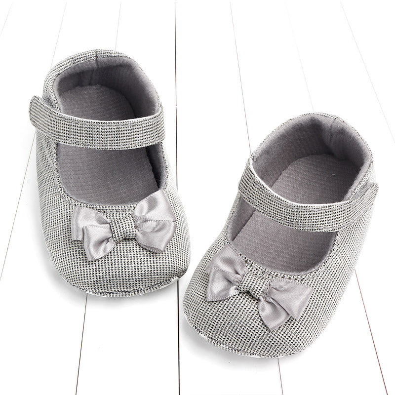 Gray baby shoes with a textured pattern and Velcro straps are adorned with small satin bows. They rest on a white, wooden surface.