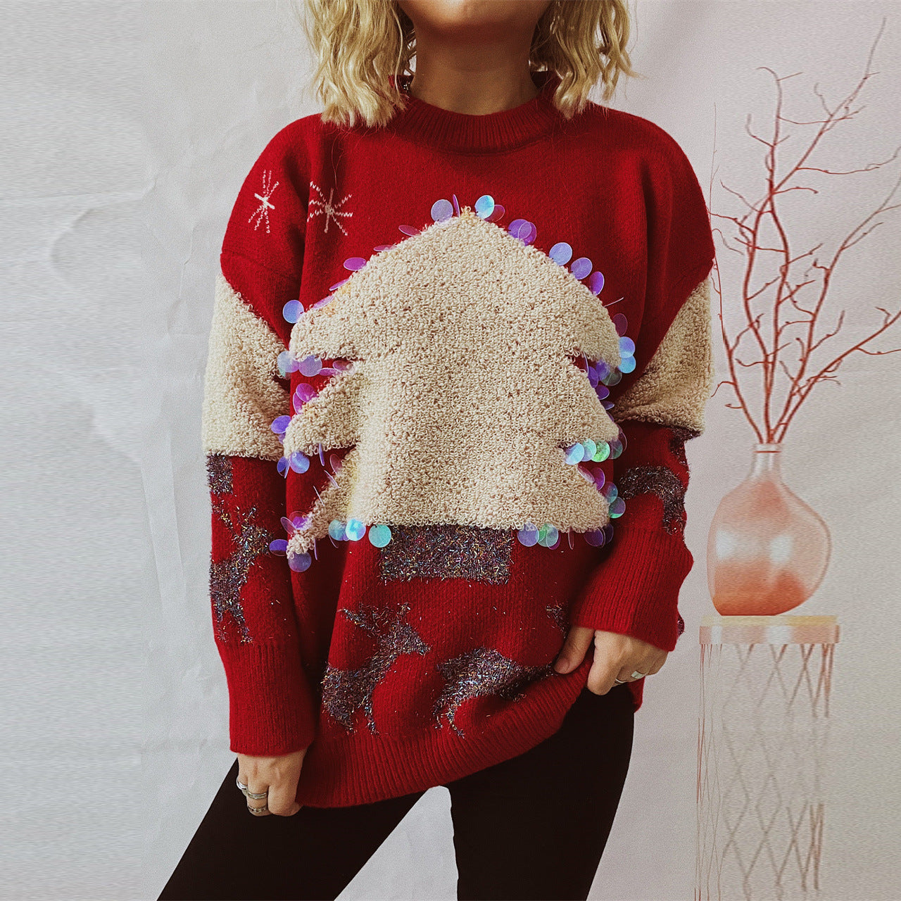 A person wears a red festive sweater featuring a textured Christmas tree adorned with sequins, alongside reindeer patterns, standing against a pale backdrop with a decorative vase.