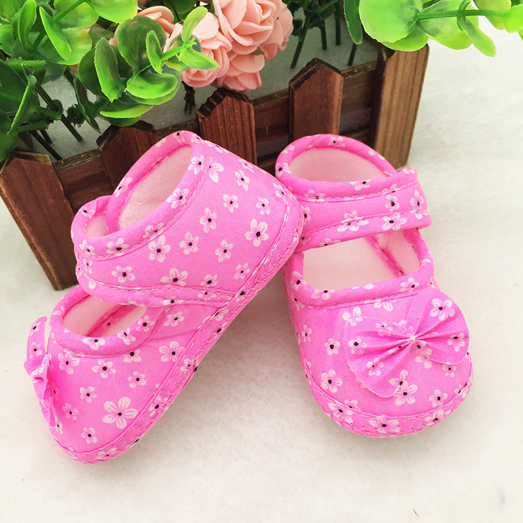Pink baby shoes with a floral pattern and bow detail, set against a background of green leaves and pink flowers in a wooden fence.