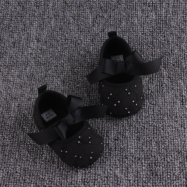 Black baby shoes adorned with small white beads and large ribbon bows, placed on a textured gray carpet.