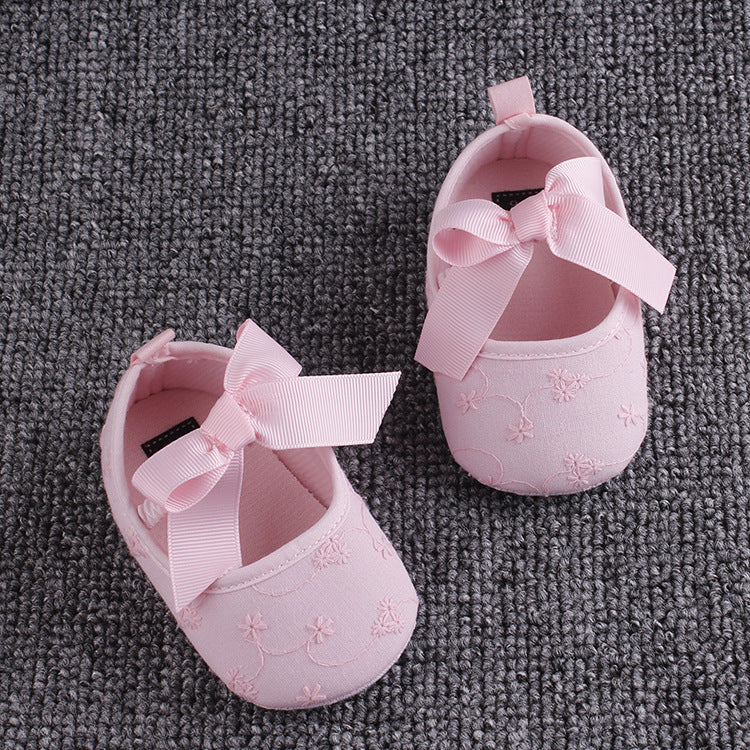 Pink baby shoes with floral embroidery and decorative bows on top, placed on a textured gray carpet.