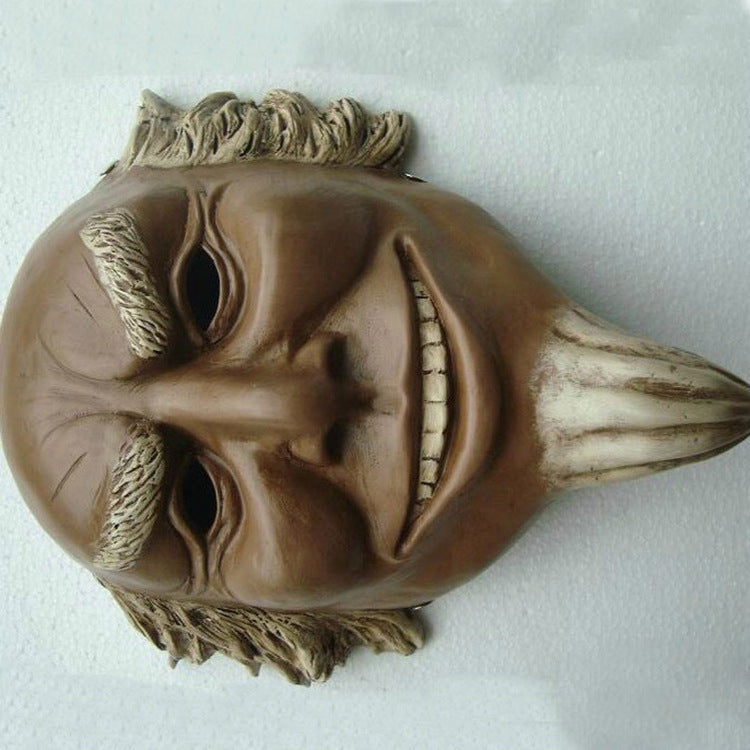 A clay mask with exaggerated features, including bushy eyebrows, a wide grin, and a pointed chin, lies on a white surface. The mask has textured hair and a smooth finish.
