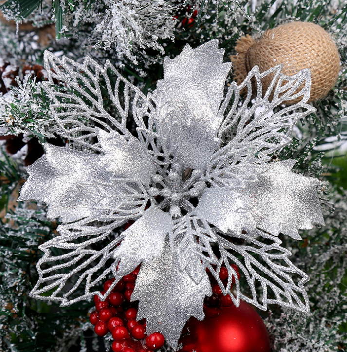 Silver glittering poinsettia ornament on a snow-dusted Christmas tree, adorned with red berries and a burlap-wrapped bauble.