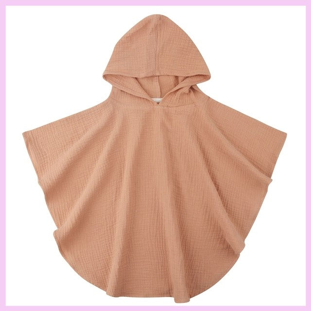 Beige hooded poncho with a relaxed fit, draping fabric, and lightweight texture. It features a soft, rounded hem and is displayed against a light pink frame background.