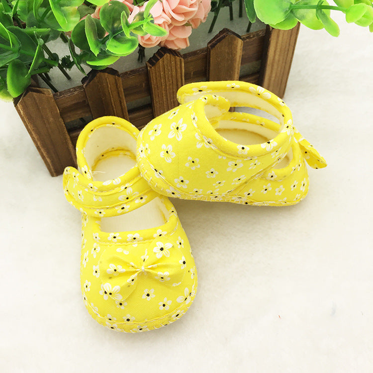 Bright yellow baby shoes with dainty white floral pattern and bow detail, displayed on a soft white surface. Green leaves and pink flowers in a wooden planter are in the background.