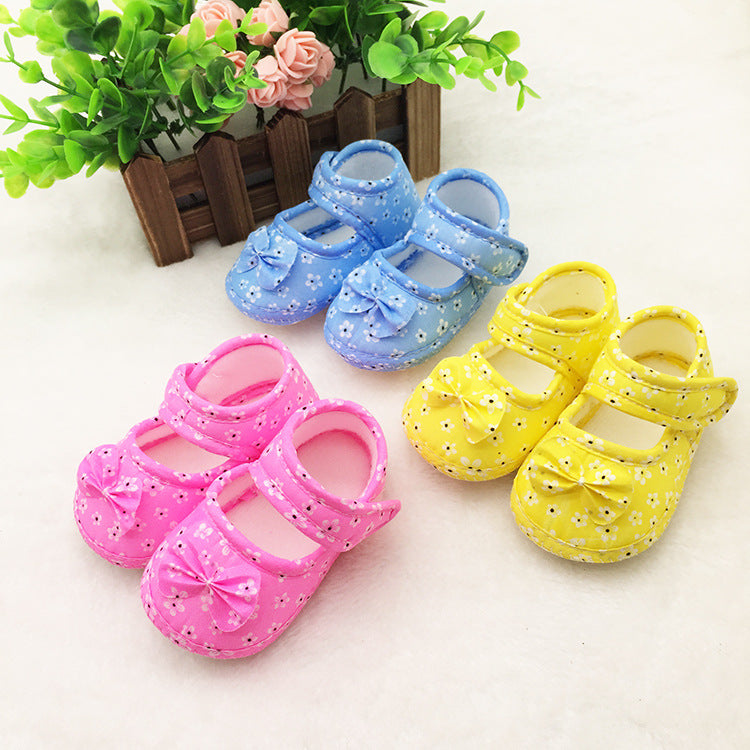 Three pairs of baby shoes in pink, blue, and yellow with floral patterns and bows are arranged on a soft white surface with a small basket of green leaves and pink flowers in the background.